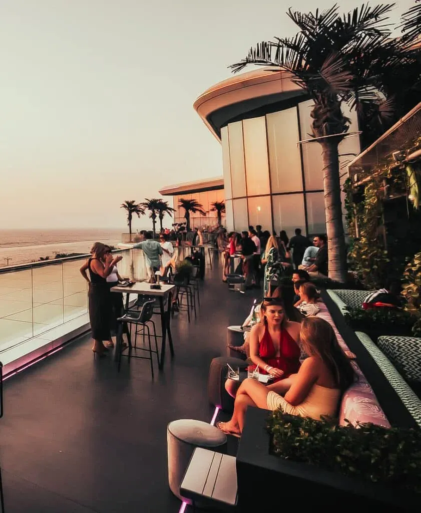 People enjoying drinks at a stylish rooftop bar at sunset, with tables and lounge seating, palm trees, and a view of the ocean in the background.