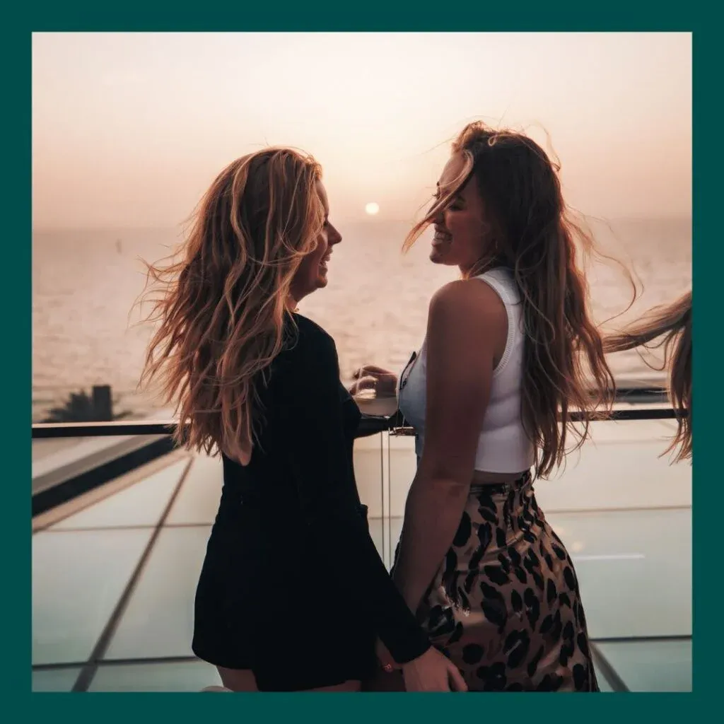 Two women with long hair standing on a balcony at sunset, smiling and looking at each other, with the ocean in the background.