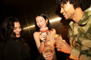 Three people are smiling and holding drinks in a dimly lit setting, suggesting a social gathering or celebration.