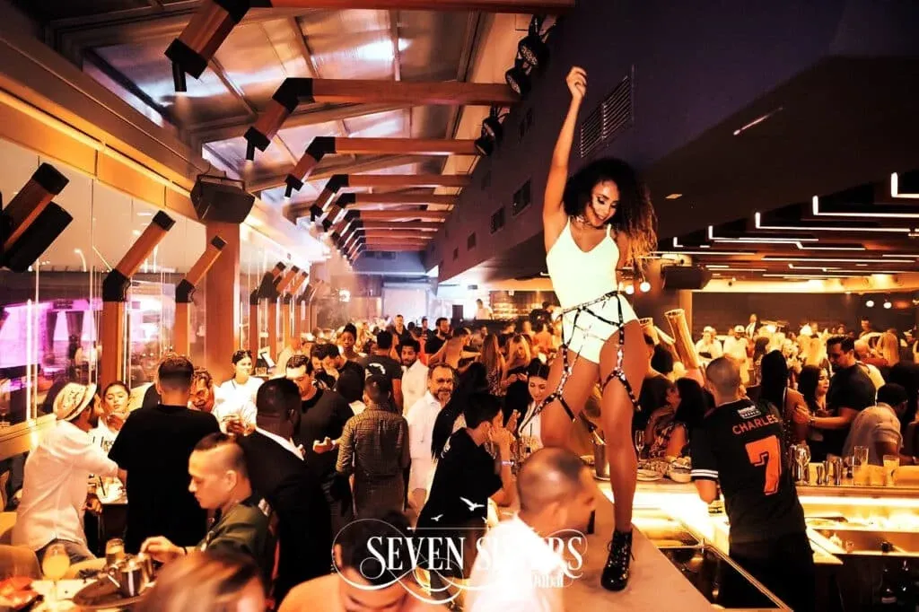 Crowded nightclub scene with people socializing; a woman is dancing on a bar in the foreground, wearing a white outfit and black boots. The location is identified as 'Seven Sisters Dubai' by text in the image.