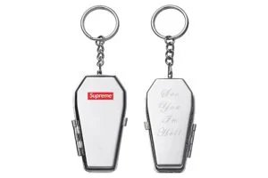 Two silver coffin-shaped keychains with hinged lids; one features a red logo, while the other is engraved with 'See You In Hell.'