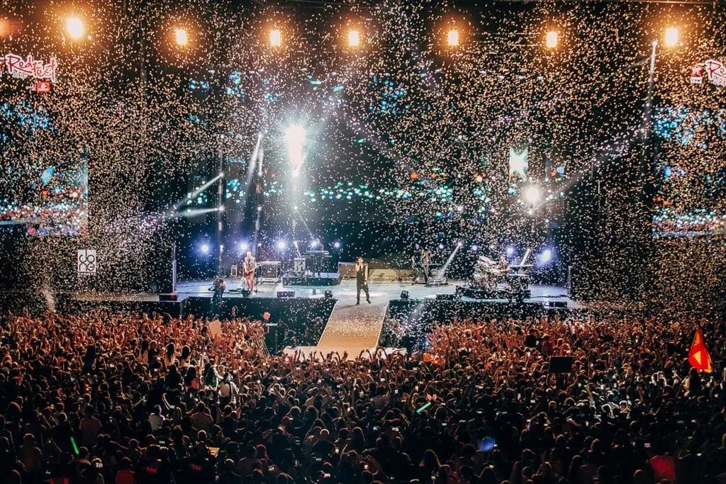 A crowded concert with a band performing on stage, bright lights, and confetti falling over the cheering audience.