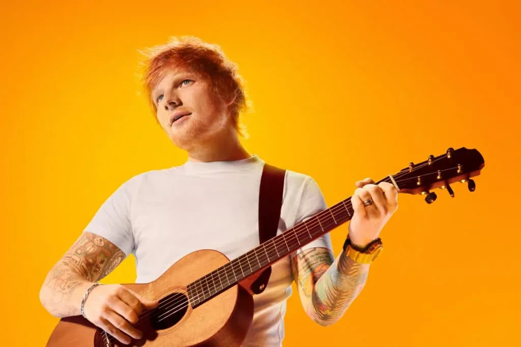 A person with ginger hair and tattoos on their arms is playing an acoustic guitar against a bright orange background.