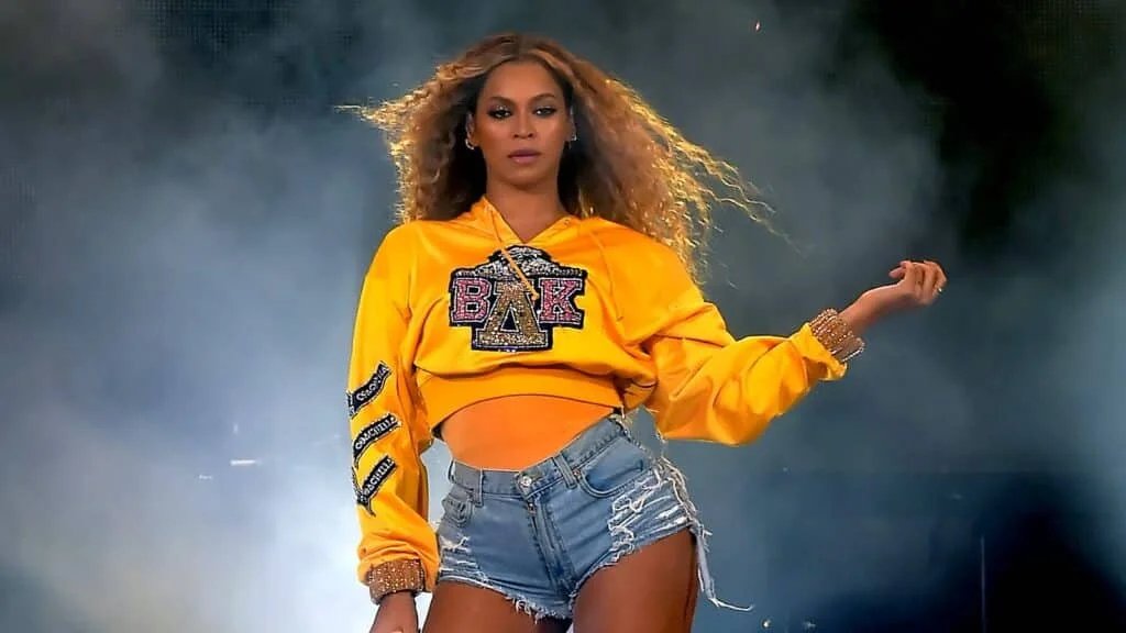 A woman performing on stage wearing a bright yellow hooded sweatshirt with Greek letters and distressed denim shorts, with flowing hair and a dramatic stage lighting backdrop.
