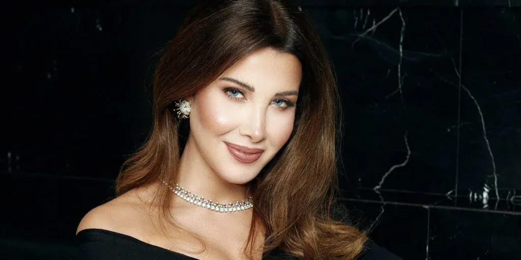A woman with long brown hair, wearing a black off-shoulder top, a pearl and diamond necklace, and floral earrings, smiling at the camera against a dark marbled background.