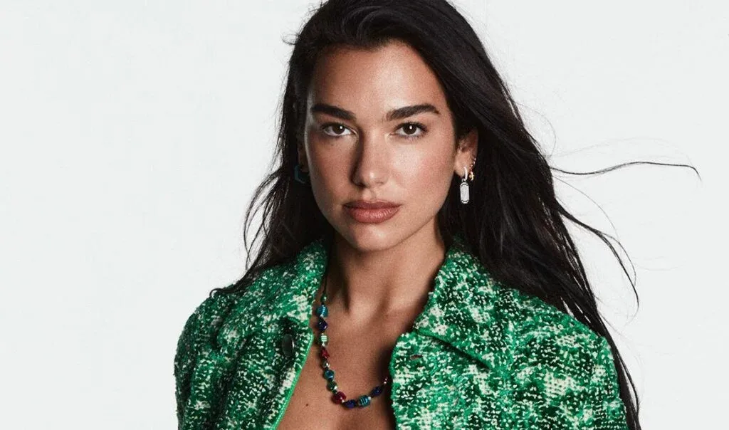 A woman with long dark hair wearing a textured green jacket and a colorful beaded necklace, standing against a plain background.