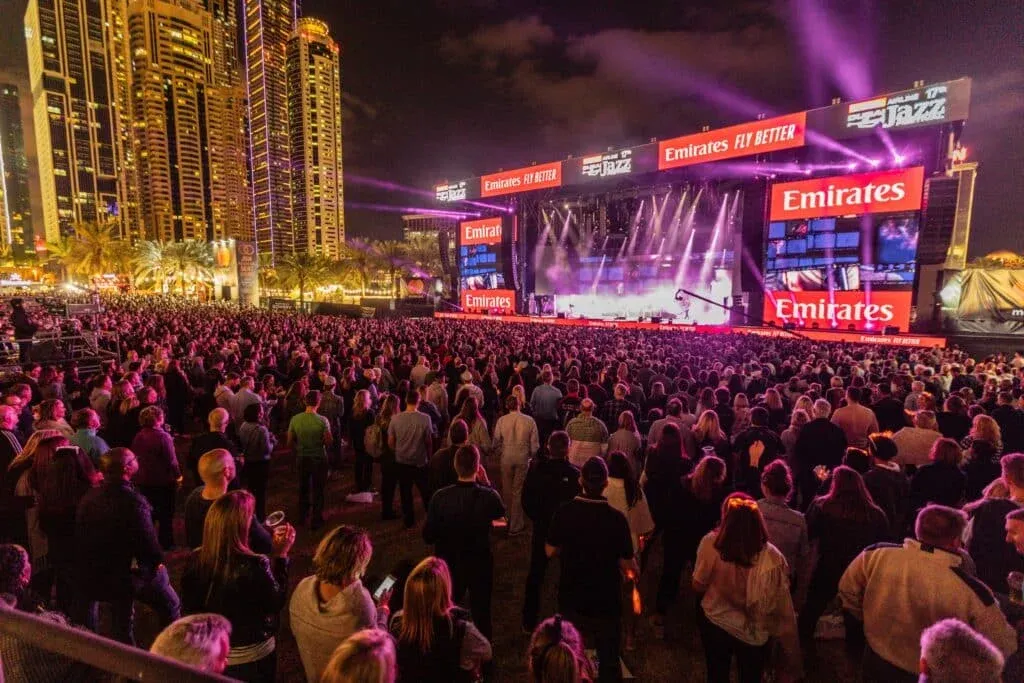 A large crowd gathers at an outdoor music concert at night, with a brightly lit stage and towering skyscrapers in the background. The event appears to be sponsored by Emirates, as indicated by prominent signage.