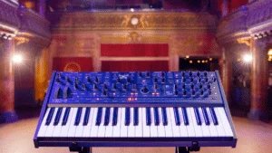 A synthesizer on a stand in a dimly lit theater with ornate details and a red velvet curtain in the background.