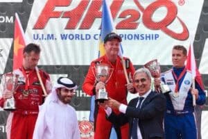 Podium ceremony at the F1H2O UIM World Championship with three racers holding trophies while standing on the podium. The first-place winner is holding a trophy and flag, with others standing beside them. Officials in suits are presenting the awards.