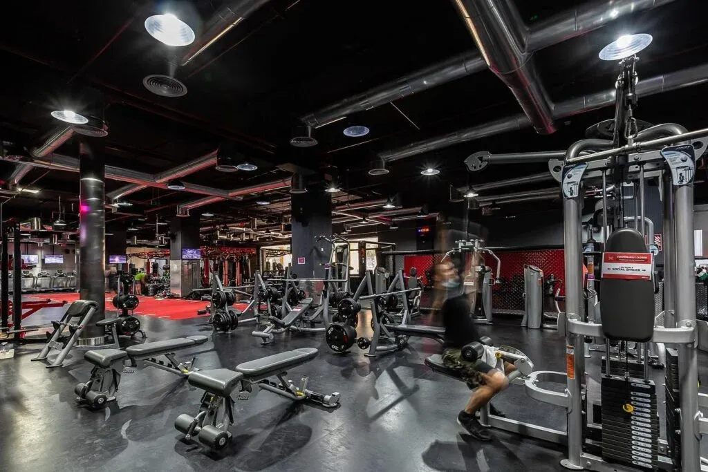 A modern gym interior with various exercise machines and benches, a person using a weight machine, red accents, and industrial-style ceiling lights and ductwork.