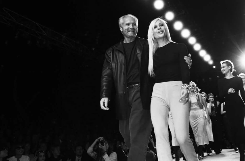 A black and white photo of a man and a woman walking arm in arm on a fashion runway, surrounded by models and bright stage lights.