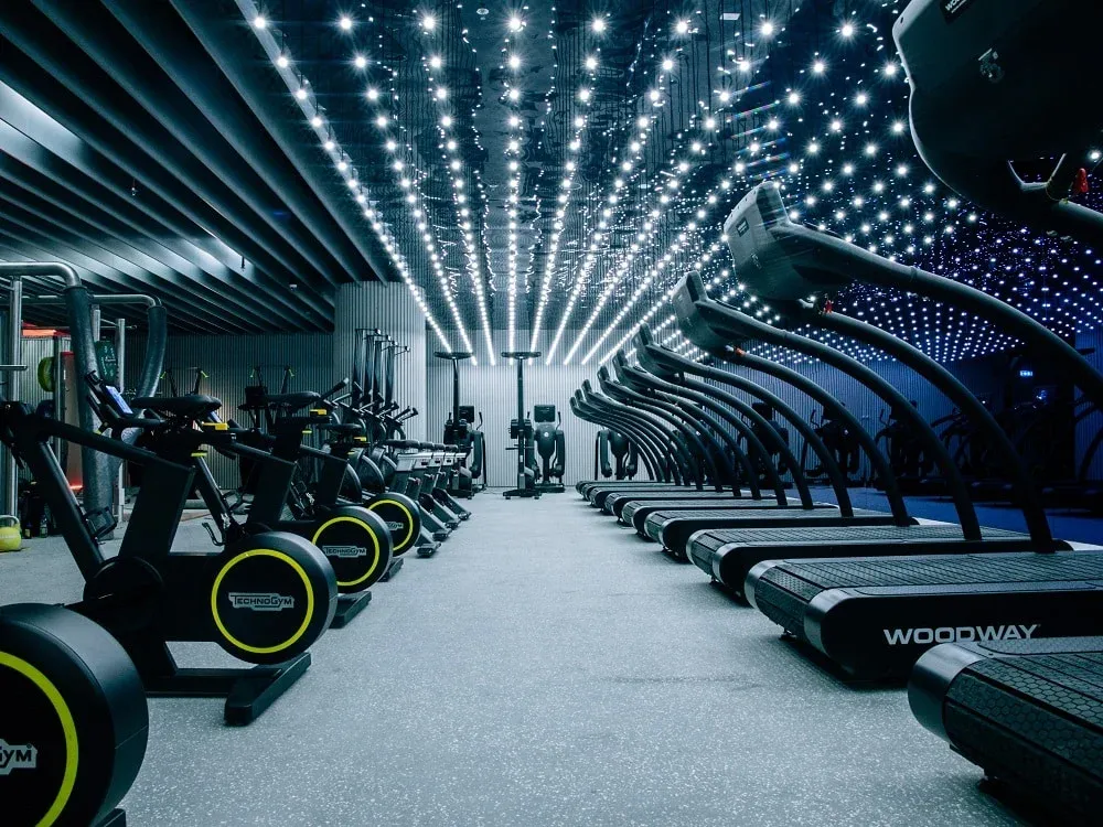 A modern gym interior featuring rows of treadmills and exercise bikes under a ceiling with numerous small, bright lights, creating a futuristic ambience.