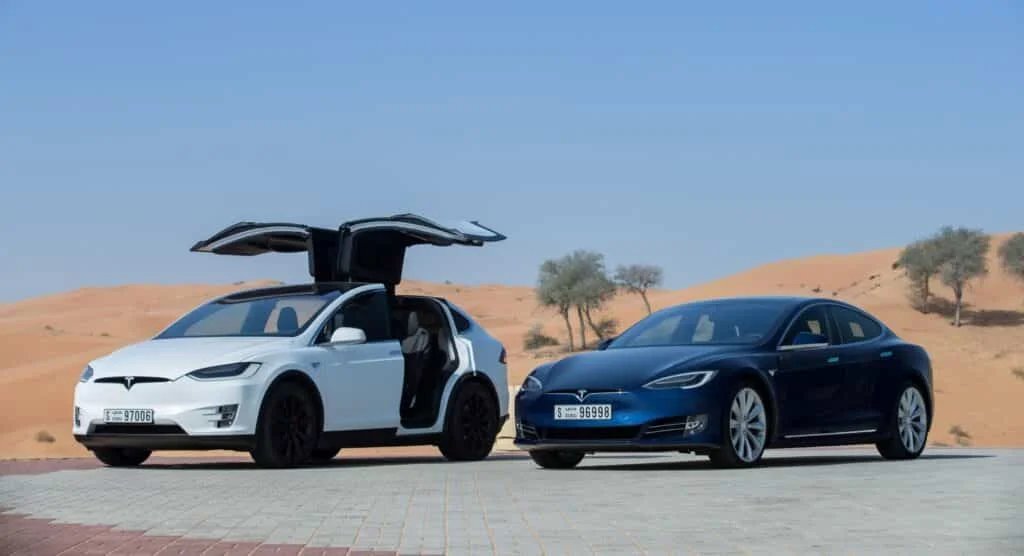 Two Tesla cars parked on a paved area in a desert, with the white Tesla Model X having its falcon-wing doors open, and a blue Tesla Model S beside it, under a clear blue sky.