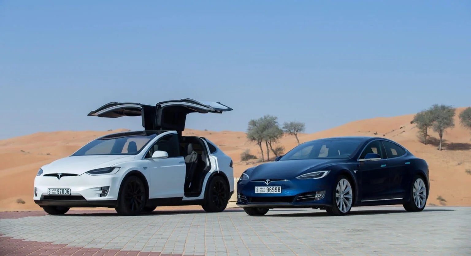 Two Tesla cars parked on a paved area in a desert, with the white Tesla Model X having its falcon-wing doors open, and a blue Tesla Model S beside it, under a clear blue sky.