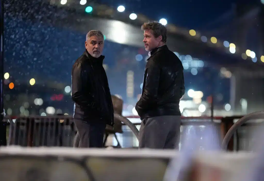 Two men in dark jackets standing outside at night, with blurred city lights and a bridge in the background.