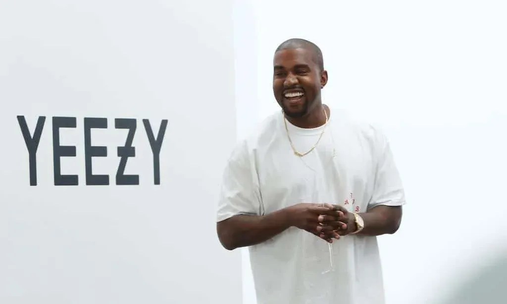 A person smiling broadly wearing a white t-shirt and gold jewelry, standing in front of a wall with the word 'YEEZY' in large, bold letters.