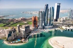 Aerial view of a modern cityscape with high-rise buildings along a waterfront. The skyline features several tall, uniquely designed skyscrapers, surrounded by green parks and sandy beaches adjacent to a blue body of water.