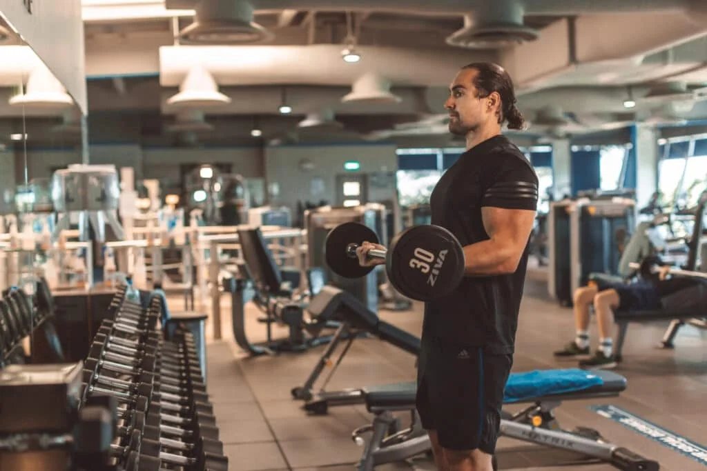 A person in athletic clothing doing bicep curls with a dumbbell in a gym, surrounded by weights and exercise equipment.