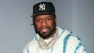 A man wearing a black baseball cap with a white logo, a light blue and white tie-dye hoodie, and multiple gold chains, against a plain background.