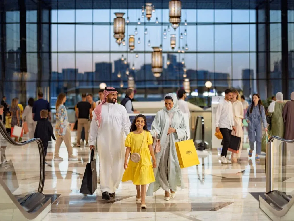 A family with a father, mother, and daughter walking through a spacious shopping mall with large glass windows. The father is wearing traditional attire and holding a black shopping bag, the mother is wearing a light blue outfit and carrying a yellow Fendi bag, and the daughter is dressed in a yellow dress with a small purse. Other shoppers are visible in the background.