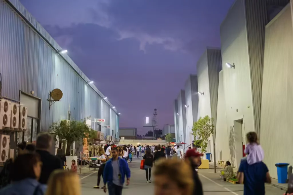 The image shows a busy outdoor scene in an industrial area at dusk. Tall warehouse-like buildings with corrugated metal walls line both sides of the walkway, illuminated by outdoor lights. A crowd of people, including a person carrying a child on their shoulders, are walking along the path, creating a bustling atmosphere under the twilight sky.