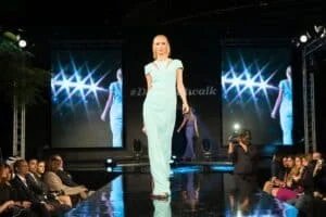 A model wearing a light blue dress walks down a glossy runway at a fashion show, with an audience seated on both sides and a cameraman capturing the moment. Two screens in the background display a reflection of the model in a similar pose.