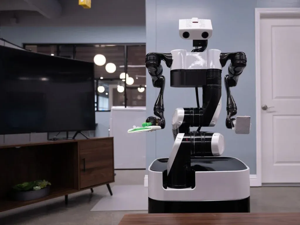 A robot with black arms and a white body is holding a small green object in a room with modern decor, including a flat-screen TV and a wooden cabinet.