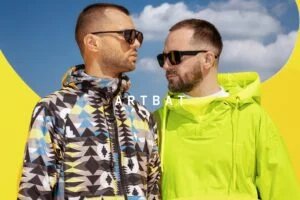 Two men wearing sunglasses, one in a colorful patterned jacket and the other in a bright green hooded jacket, standing under a blue sky with clouds and a yellow backdrop, with the text 'ARTBAT' across the image.