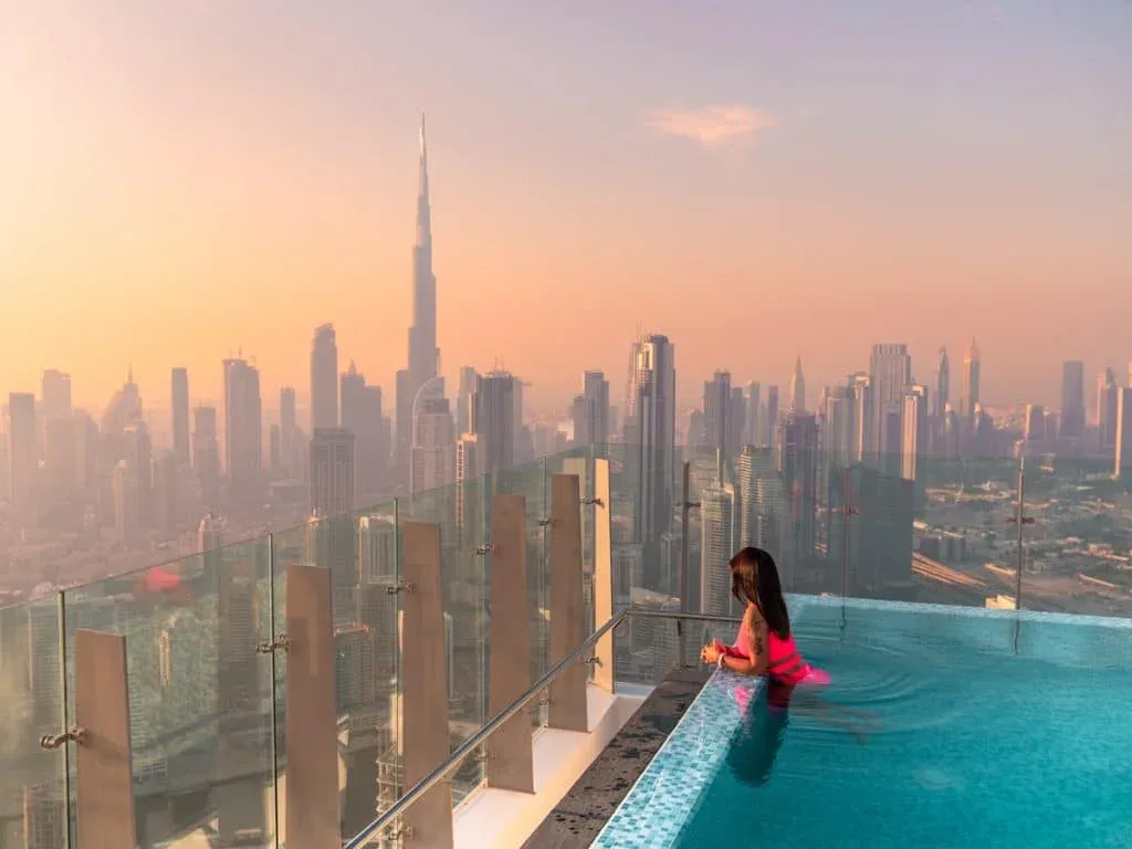 A woman in a pink swimsuit stands in an infinity pool overlooking the Dubai skyline, with the Burj Khalifa visible amid a hazy sunset sky.