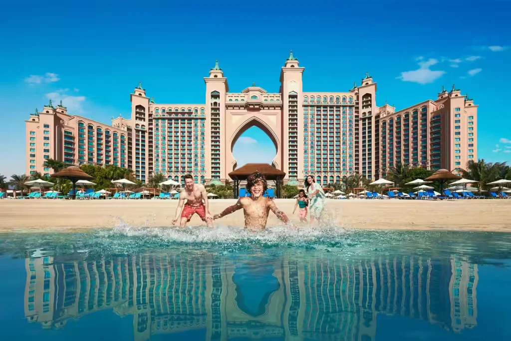 A group of people enjoying the beach in front of a large, luxurious hotel with an arched design. The scene includes blue skies, umbrellas, and sun loungers, with reflections of the building visible in the water.