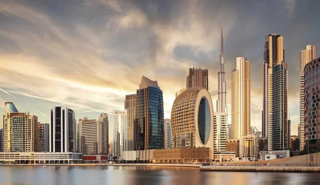 A cityscape of Dubai featuring modern skyscrapers including the Burj Khalifa amidst a vibrant sunset sky, with reflections on a body of water in the foreground.