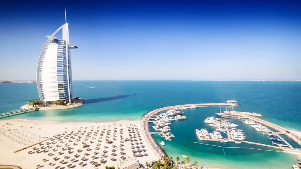 Aerial view of Burj Al Arab hotel on a man-made island, surrounded by turquoise water and adjacent to a marina with yachts. A sandy beach with rows of umbrellas stretches along the shoreline.