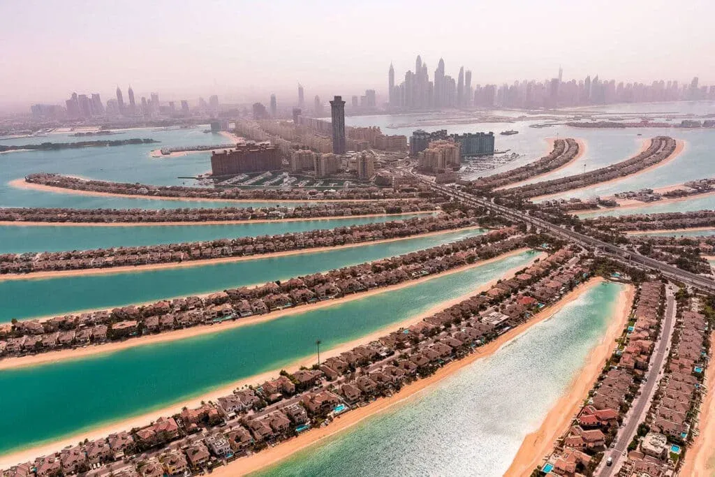 Aerial view of the Palm Jumeirah in Dubai, featuring a man-made palm-shaped island with residential villas, surrounded by turquoise water, and a modern skyline in the distant background.