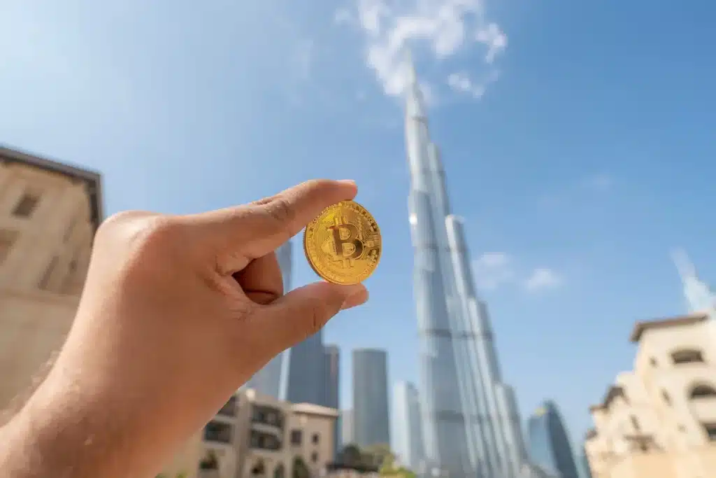 A hand holding a gold Bitcoin coin with the towering Burj Khalifa skyscraper blurred in the background against a clear blue sky.