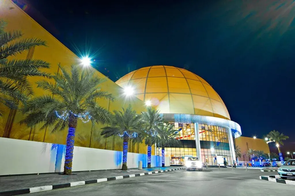 A large, illuminated dome-shaped building at night, with palm trees wrapped in blue lights, and a parked vehicle in front.