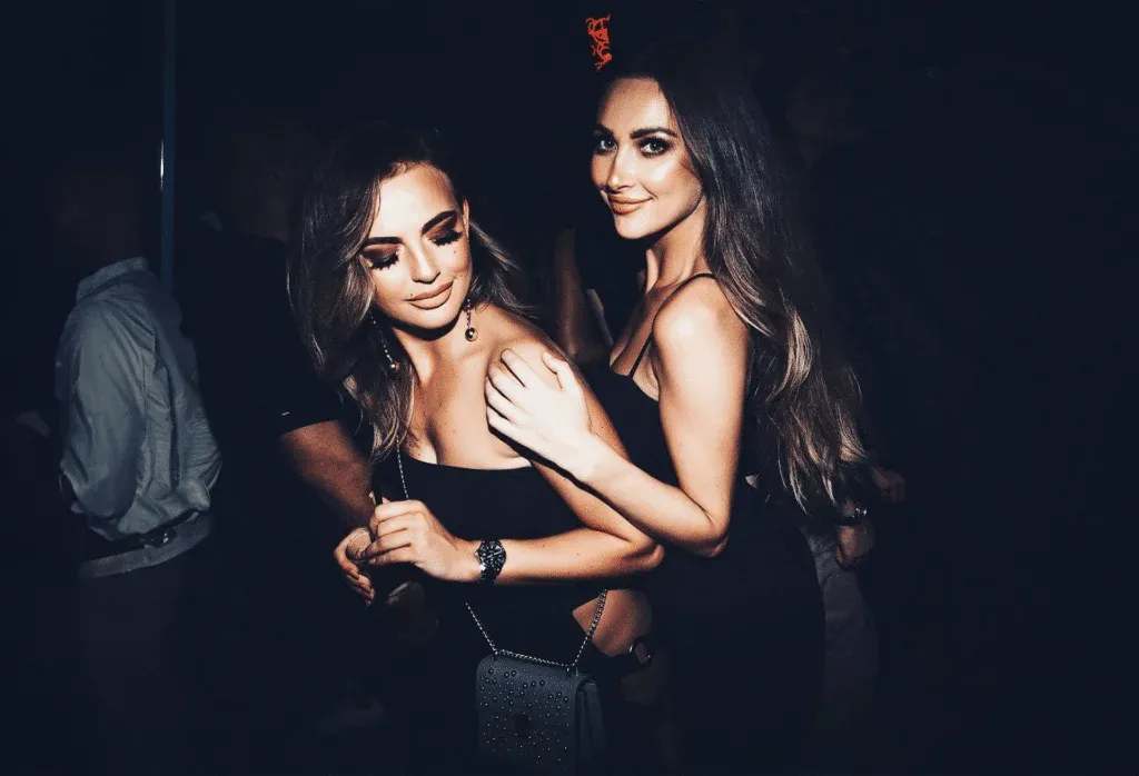 Two women in black dresses are smiling and posing together in a dimly lit environment.