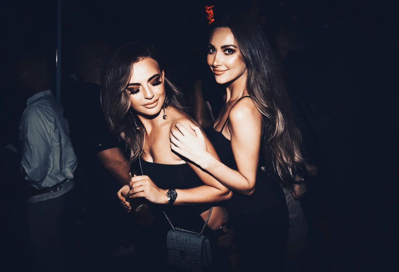 Two women in black dresses are smiling and posing together in a dimly lit environment.