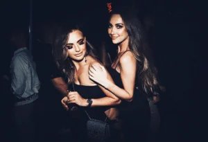 Two women in black dresses are smiling and posing together in a dimly lit environment.