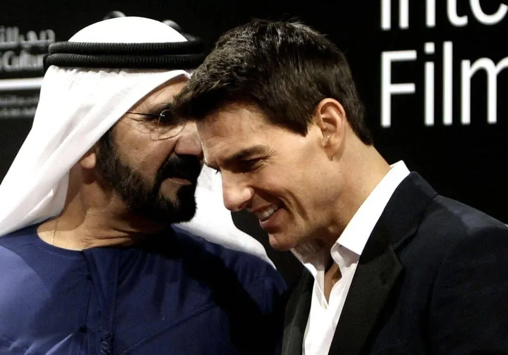 Two men on a stage, one wearing a traditional white headscarf and black suit, conversing closely and smiling.