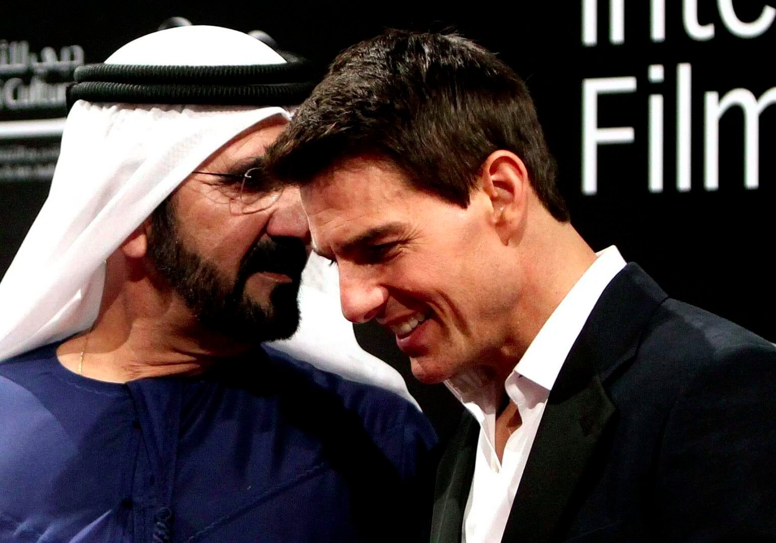 Two men on a stage, one wearing a traditional white headscarf and black suit, conversing closely and smiling.