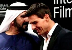 Two men on a stage, one wearing a traditional white headscarf and black suit, conversing closely and smiling.