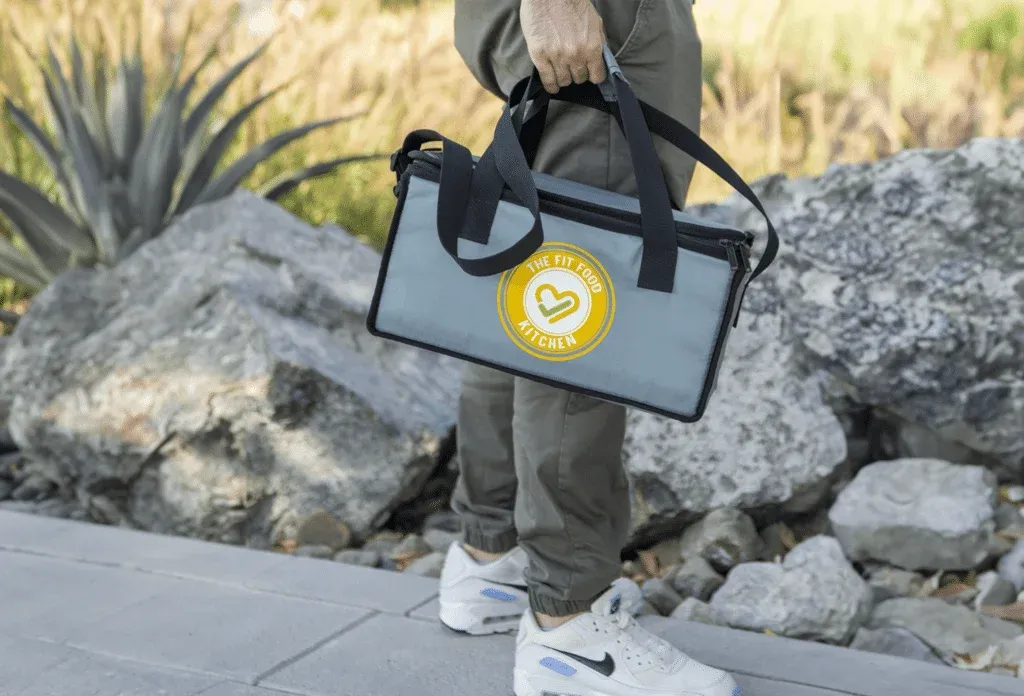 A person holding a gray insulated bag with 'The Fit Food Kitchen' logo, standing on a paved path with rocks and plants in the background.