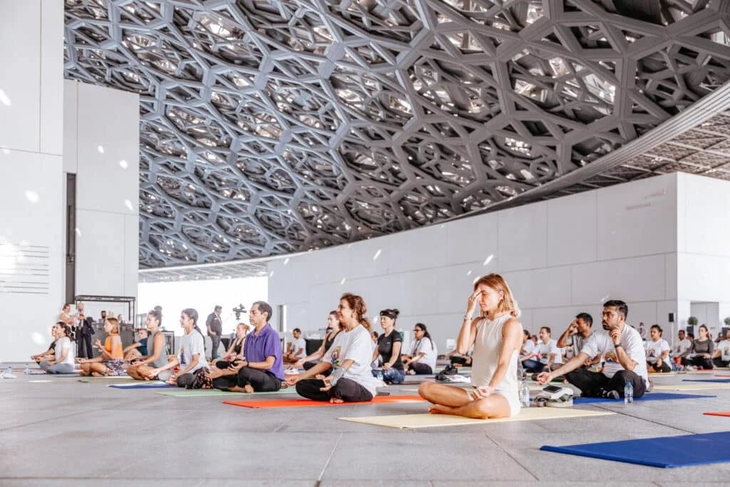 A group of people sitting cross-legged on yoga mats in a large, architecturally elaborate indoor space, practicing meditation or breathing exercises.