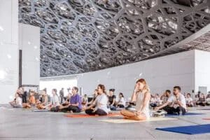 A group of people sitting cross-legged on yoga mats in a large, architecturally elaborate indoor space, practicing meditation or breathing exercises.