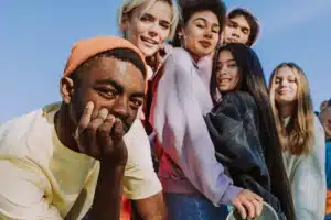 A group of young adults smiling and posing outdoors against a clear blue sky; the person in the foreground wears a peach-colored beanie and rests their chin on their hand.