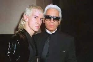 Two people posing together; one with mullet-style blond hair and wearing a shiny black jacket, the other with short white hair, wearing sunglasses and a black suit with a high collar.