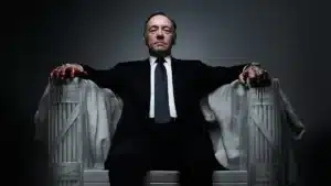 A man in a suit sits on a large chair with his arms resting on the armrests, hands stained with dripping blood.