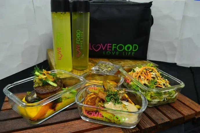 A collection of glass containers holding assorted gourmet dishes, including grilled vegetables and salads, arranged on a wooden board. Two bottles labeled 'Love Food Love Life' contain a yellow beverage, and a black insulated bag with the same logo is in the background.