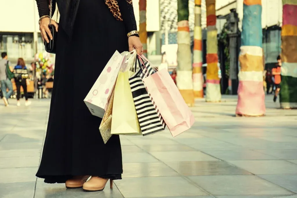 A person wearing a long black dress holding several colorful shopping bags in an outdoor shopping area with artistically wrapped columns in the background.