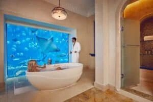 A woman relaxes in a bathtub inside a luxurious bathroom with a large aquarium window featuring rays and fish. A man in a white bathrobe stands nearby, holding a drink.